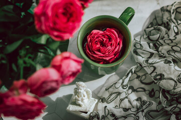A green ceramic mug, in which a blossoming red rose bud is floating, stands on a gray table, next to a vase with a bouquet of scarlet roses; shot from above