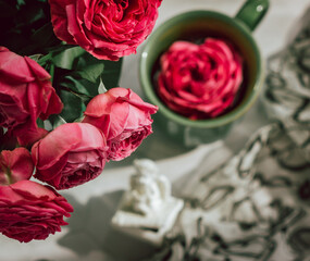 A green ceramic mug, in which a blossoming red rose bud is floating, stands on a gray table, next to a vase with a bouquet of scarlet roses; shot from above