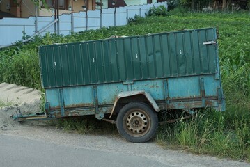 one old metal trailer on wheels stands on the gray asphalt of the road near the grass on the street