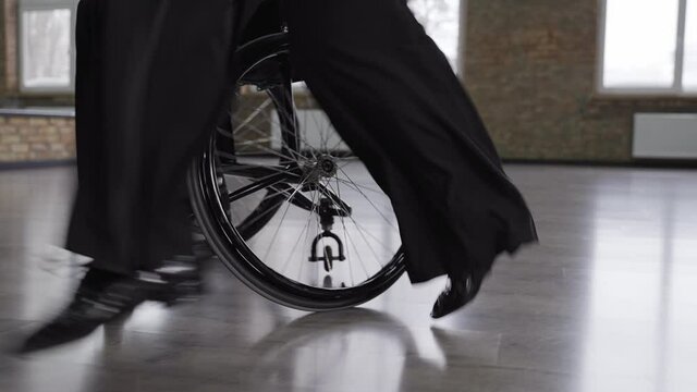 Close-up of male dancer legs skillfully making dance pas next to female partner sitting on wheelchair. Low section of professional ball dancers in black clothing performing active dance moves