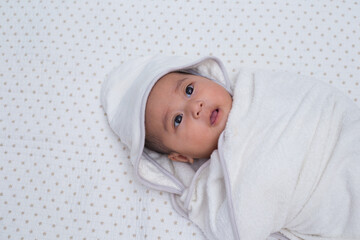 Baby girl wrapped in white towel after bath, lying on bed. Southeast asian 4 months old daughter on white background wrapped, looking upward. 