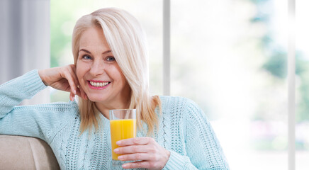 Happy woman holds a glass of orange juice and looks relaxed at the camera, sitting near the window.
