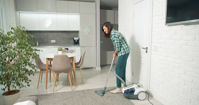 Young woman cleaning dust and debris from gray carpet with vacuum cleaner in living room at home.