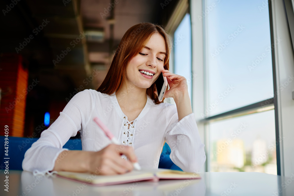 Aufkleber cheerful female smiling while writing informative notes during positive cellphone conversation for d