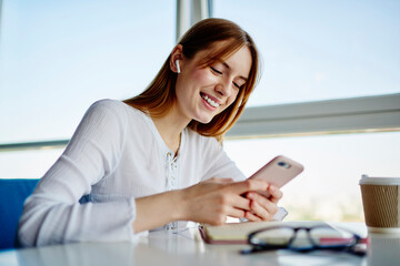 Funny female student in electronic earbuds enjoying mobile messaging in social networks using...