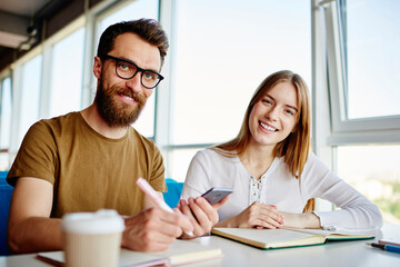 Obraz premium Portrait of cheerful male and female students smiling at camera during together learning in campus, happy Caucasian bloggers posing during daytime meeting for discussing mobile phoning via app