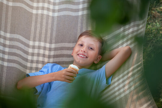 Cute Smiling Caucasian Boy Eating Ice Cream And Having Fun On Hammock In Backyard Or Outdoor.Child Staying Cool In The Summer Heat.