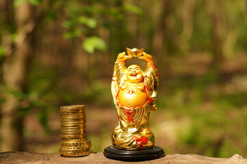 A figure of a happy smiling Buddha on a colored natural background. There is a stack of coins next...