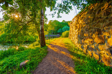 Naklejka premium Alley going between the fortress and the river. The sun shines through the foliage of the trees. Beautiful nature. Priozersk, Russia. View from the road near the fortress wall.