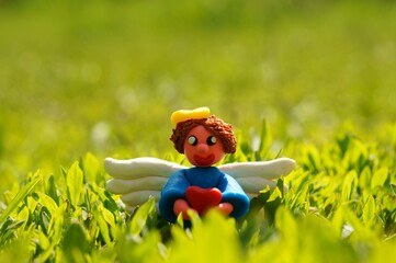 A figurine of an angel made of plasticine with a heart in his hands on a background of green grass.