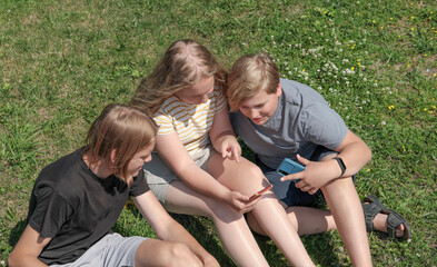 group of teenagers looking at smartphone screen and smiling and chatting. teens sitting on a lawn grass wearing casual clothes spending time outdoors