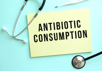 The text ANTIBIOTIC CONSUMPTION is written in a yellow pad that lies next to the stethoscope on a blue background.