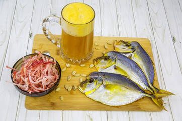 A mug of beer with foam, dried vomer fish and squid shavings in a round black plate on a wooden board with a wooden table background. Food is a delicacy snack for beer.