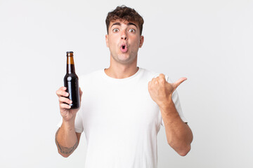 young handsome man looking astonished in disbelief and holding a beer bottle
