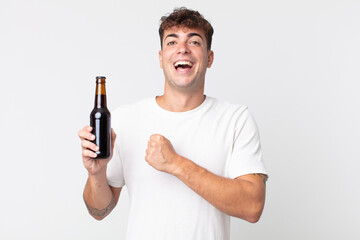 young handsome man feeling happy and facing a challenge or celebrating and holding a beer bottle