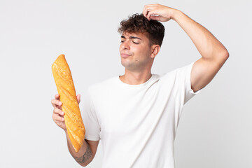 young handsome man feeling puzzled and confused, scratching head and holding a bread baguette