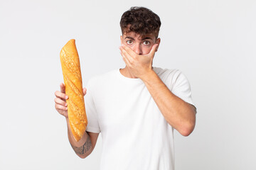 young handsome man covering mouth with hands with a shocked and holding a bread baguette