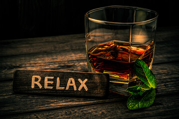 Glass of brandy with mint sprig and the wooden plank on it is an inscription "RELAX" on an old wooden table. Angle view, focus on the inscription