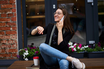 Happy hipster girl in classic spectacles smiling during positive cell talking in roaming, joyful female checking time on wearable smartwatch while making international friendly conversation via app