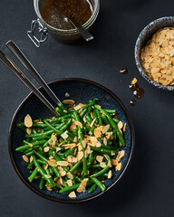 Green beans in a black bowl on a gray table. Top view. There is a spoon in a bowl. Jars with sauce and almonds stand near. Close up