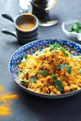 Indian Pilaf with Cauliflower yellow coloured in a blue plate on a gray table. 45 angle view. There are bowls of herbs and sprinkled seasoning on the table