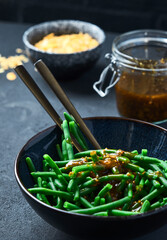 Green beans in a black bowl on a gray table. 45 view. There is a spoon in a bowl. Jars with sauce and almonds are on the background. Close up. 45 angle view