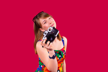 Smiling pretty young blonde woman in multicolored dress having sunglasses on the head and a bracelet on the hand holding the retro photo camera.