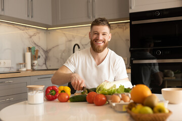 Fototapeta premium Young man sit at table and cut cucumber on salad
