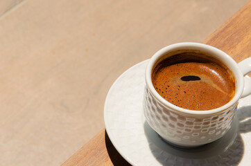 White cup of coffee with saucer stands on table on beige background. Time for coffee background with copy space. Mug with hot drink in the sun.