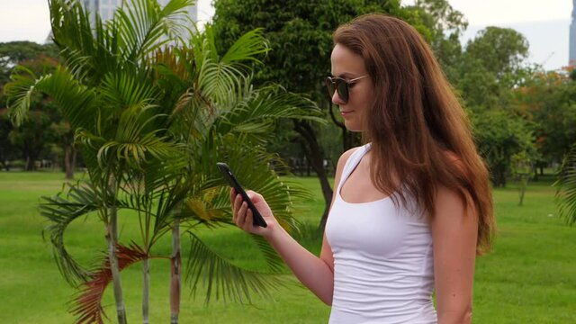 Long hair of beautiful lady fly on wind, she stand still and stare to phone, slow motion shot. Tourist recreate at scenic Lumphini park, green palm fronds sway on wind, lush vegetation on background