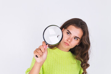 Young beautiful woman with freckles light makeup in sweater on white background with magnifier searching focused