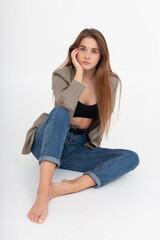 portrait of young attractive caucasian woman with long hair in suit jacket and blue jeans isolated on white background. skinny pretty female sitting on floor at studio. model tests of beautiful lady