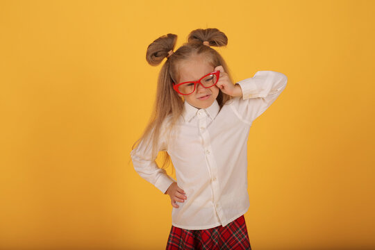 a beautiful little smiling blonde girl with red-rimmed glasses in a white shirt and a plaid skirt is a schoolgirl on a yellow background