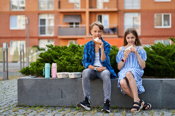 Children girl and boy sitting near school and have lunch of sandwich in pink and blue lunchbox and...