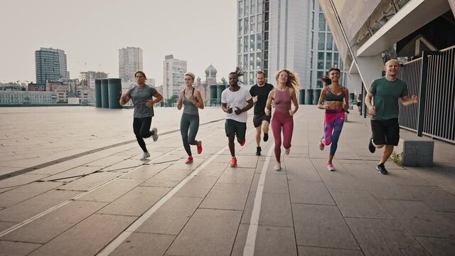 Front View. Running Club, Group Of Like-minded People Run Early In The Morning At Dawn Along The Urban Streets. Multiethnic Team Of Men And Women Training Running Before Marathon, Active Life