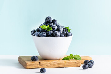 White bowl with fresh organic blueberries on light blue background close up. Healthy summer dessert on the table.