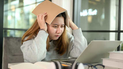 Asian teenage girl holding a book on her head stressed looking at laptop during online lessons from laptop.