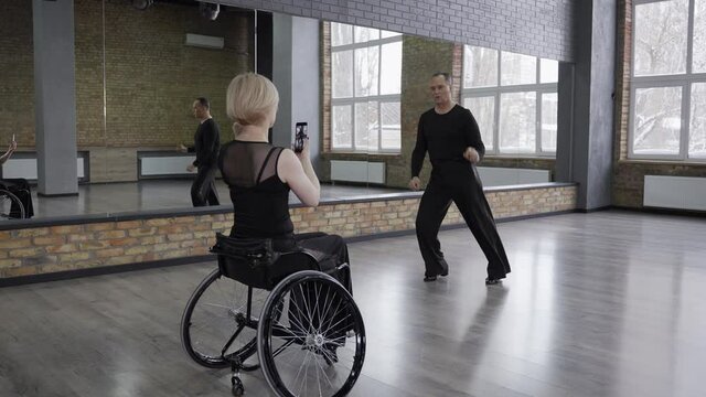 Handicapped woman on wheelchair using cellphone while taking video of man partner dancing in ballroom. Imposing ball dancer skillfully dancing on camera filmed by female partner with impaired mobility