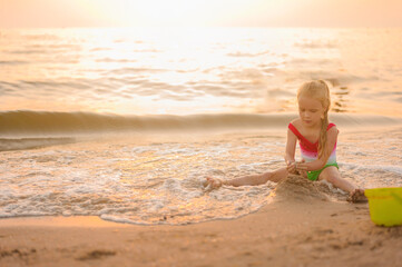 little blonde girl builds a sand castle on the seashore in the evening at sunset in the golden hour in summer