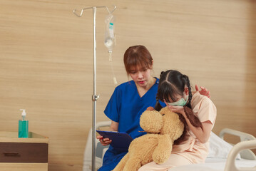 Young Asian female caregiver holding protective face mask with cute girl patient in hospital. Nurse...