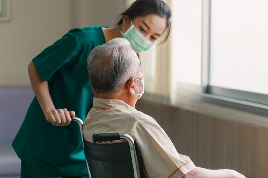 Young Asian Woman Nurse Explaining Information To Elderly Man Patient In Wheelchair With Friendly Smiley Face In The Hospital. Young Assistance With Old People In The Elderly Care Place