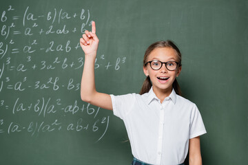 excited schoolgirl in eyeglasses showing idea gesture near chalkboard with equations