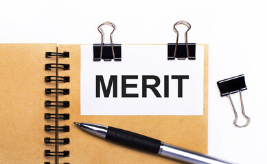 On a light background, a brown notebook with a pen, black clips and a white card with the text MERIT