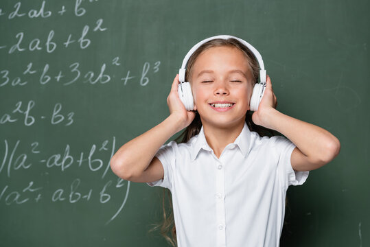 joyful schoolgirl with closed eyes listening music in headphones near chalkboard