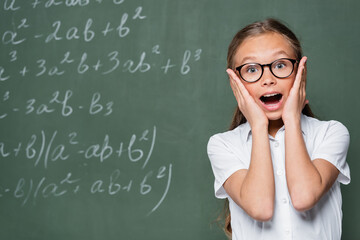 astonished schoolchild touching face and looking at camera near chalkboard