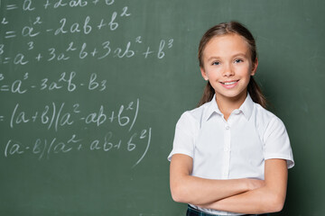 preteen schoolkid with crossed arms smiling at camera near equations on chalkboard