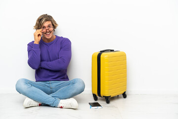 Young blonde man with suitcase sitting on the floor with glasses and happy