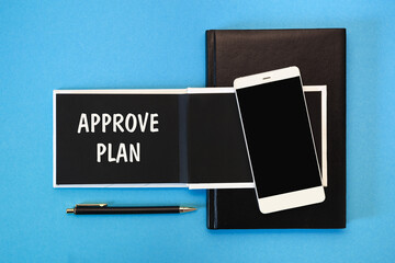 A white notebook with black pages, a smartphone and a pen on a blue background. The inscription APPROVE PLAN