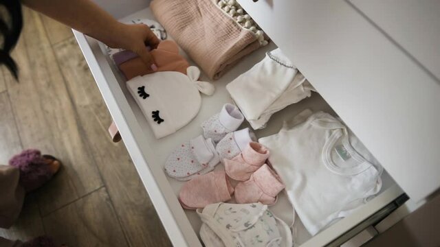 Loving parents are expecting baby and putting clothes. Children's bodysuits, hats, socks, gloves and sliders in chest of drawers. Mom and dad are waiting for newborn. Tenderness and kindness in family