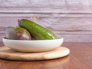 Avocados are served on a white plate on a wooden cutting board with a wood wall background. Side view. Space for text. Healthy fruits concept